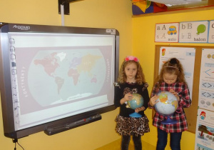Dzieci stoją pod tablicą interaktywną, trzymają w dłoniach globusy.